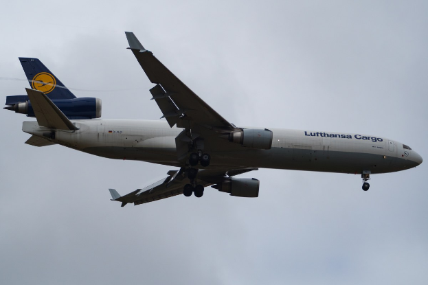 Eine Lufthansa Cargo Boeing 777-300ER im Flug, mit ausgebreiteten Tragflächen und aktivierten Triebwerken, mit sichtbarem Text auf der Rüfseite gegen einen wolkengefüllten Himmel.
