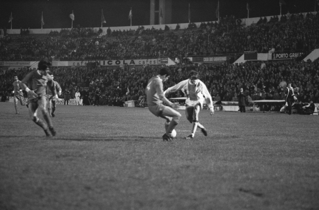 Schwarzes und weißes Bild von Männern, die Fußball auf einem Feld mit Zuschauern in den Rängen und Fahnen im Hintergrund spielen.