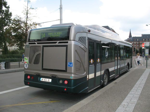 Eine Stadtbus fährt auf einer von Hochhäusern gesäumten Straße, mit Fußgängern auf einem Gehweg und Schildern auf Pfosten auf der rechten Seite und Bäumen, Gebäuden und Himmel im Hintergrund.