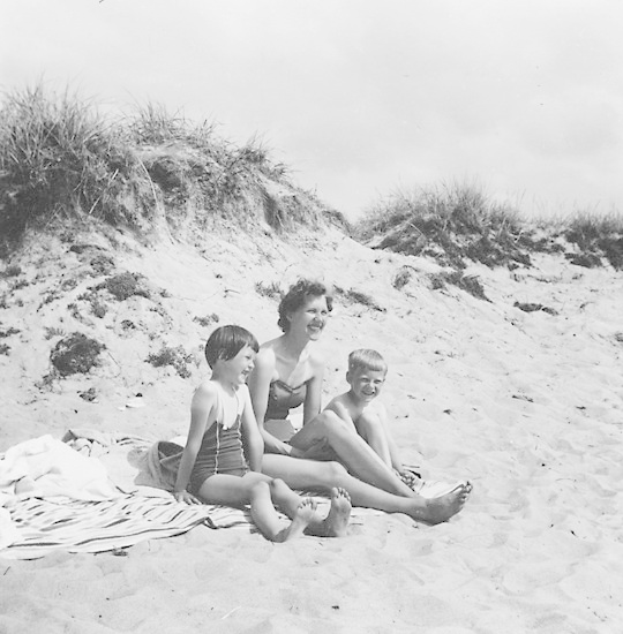 Schwarzes und weißes Foto einer Frau und zweier Kinder auf einer Stranddecke sitzend, mit Sand, Gras und Himmel im Hintergrund.