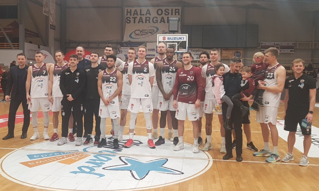 Männer- und Kinder-Gruppe auf einem Basketballfeld mit Schildern "Hala Osir Starga", sitzende Zuschauer, ein Basketballkorb, Metallstangen, Deckenbeleuchtung und eine Wand im Hintergrund.