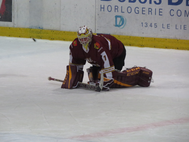 Eishockey-Spieler in rot-gelber Uniform, der einen Schuss auf dem Eis abwehrt, trägt Helm, Handschuhe und Knieschoner und hält einen Eishockey-Schläger, mit einer Wand im Hintergrund, auf der etwas geschrieben steht.