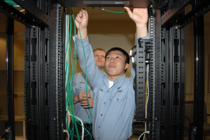 Zwei Männer arbeiten an einem Server in einem Rechenzentrum, umgeben von Kabelregalen und einer Wand im Hintergrund.