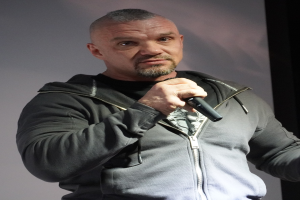 Ein Mann in einer schwarzen Kapuzenjacke und einem schwarzen Anzug mit weißem Hemd hält ein Mikrofon und steht selbstbewusst vor einer Wand, sieht aus wie ein Komiker mit zurückgekämmtem Haar.