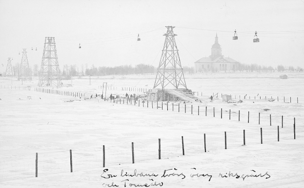 Schwarze und weiße Aufnahme eines Skilifts in einem verschneiten Feld mit Stützpfählen, Überseilbahn, Bäumen und einem Gebäude im Hintergrund, mit Text unten.