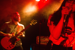 Eine Frau singt in ein Mikrofon auf der Bühne, während ein Mann Gitarre spielt und Deckenlampen die lebendige Szenerie beleuchten.