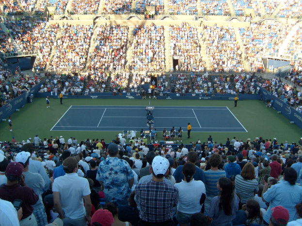 Eine große Menschenmenge schaut einem Tennisspiel in einer vollen Arena zu, mit Spielern auf dem Platz und Zuschauern in den Rängen.