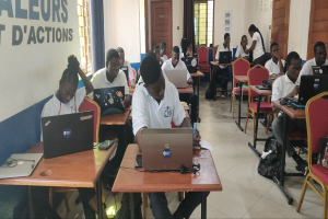 Eine Gruppe von Schülern in einem Klassenzimmer, die an Tischen mit Laptops sitzen, mit Taschen auf dem Boden und einer Wand mit Text im Hintergrund, der auf einen Online-Kurs in Ghana hinweist.