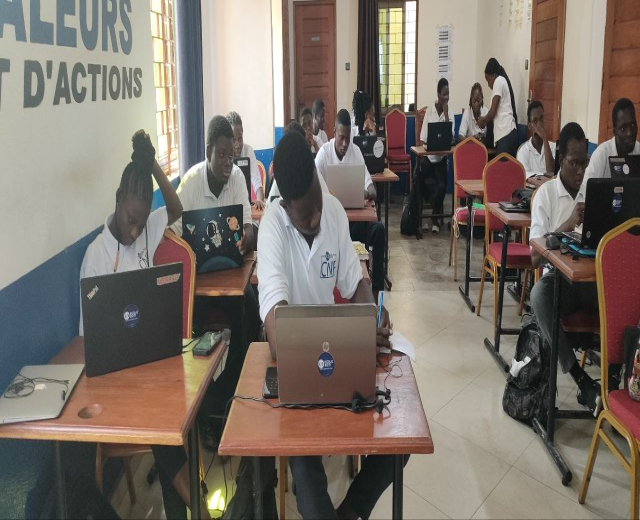 Eine Gruppe von Schülern in einem Klassenzimmer, die an Tischen mit Laptops sitzen, mit Taschen auf dem Boden und einer Wand mit Text im Hintergrund, der auf einen Online-Kurs in Ghana hinweist.