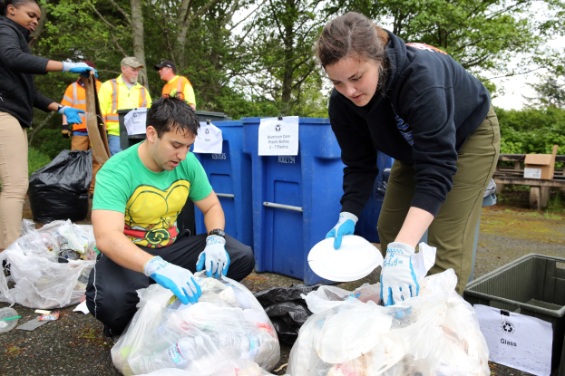 Menschen im Park Müll sammeln, darunter Plastikabdeckungen und Flaschen, mit einem Mülleimer und einer Bank im Hintergrund.