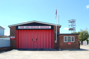 Feuerwehr- und Rettungsdienstgebäude mit roter Tür, Fenstern, einem Namensschild, einer Flagge an einer Flagge, einem Metallturm, einem Zaun, einer Baumgruppe und einem bewölkten Himmel.